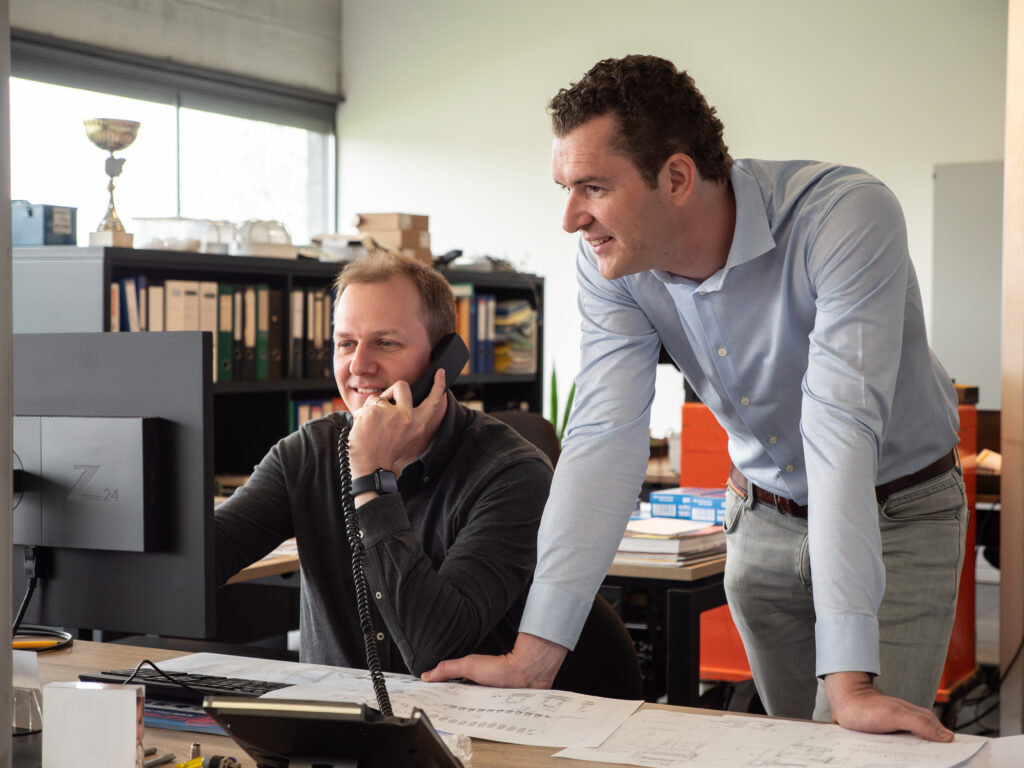 Twee Incomec-medewerkers overleggen aan een computer en bekijken technische plannen op kantoor.